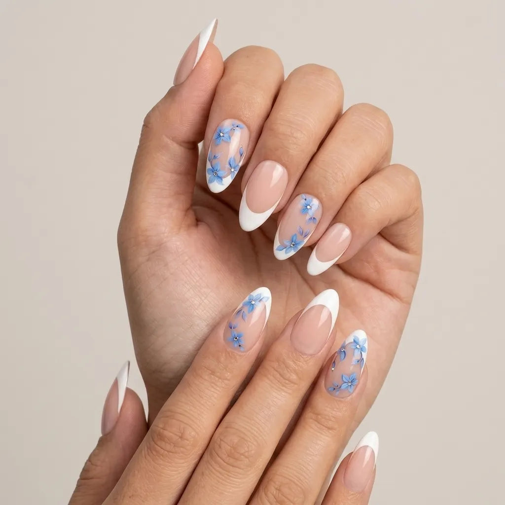A set of false press-on nails displayed in a clear plastic case, featuring a nude pink and white French tip design with blue floral accents on two nails. The case is styled on a beige surface with decorative blue flowers, a green stem, and a gold ring.
