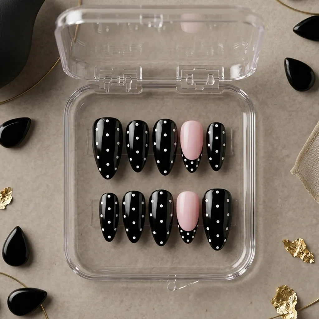 A set of black press-on nails with white polka dots and one pale pink accent nail, displayed in a clear plastic case on a neutral beige surface with scattered decorative black and gold leaf pieces.