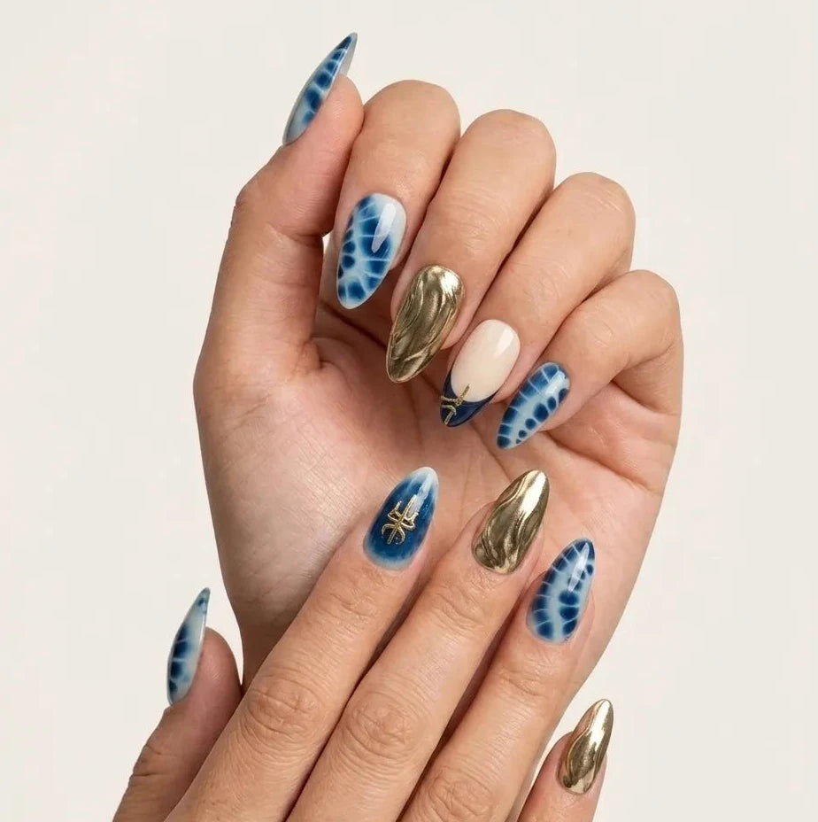 A close-up of a hand with long stiletto-shaped nails featuring a mix of metallic gold, blue-and-white tie-dye, and solid cream nail art, set against a plain light beige background.Handmade reusable press on nails
