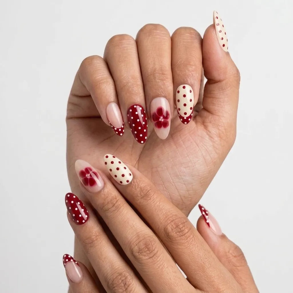 A set of 10 press-on nails displayed in a clear plastic case on a textured beige surface, decorated with red and white polka dots, floral designs, and solid pale pink tips, accented by ribbons and pearls.