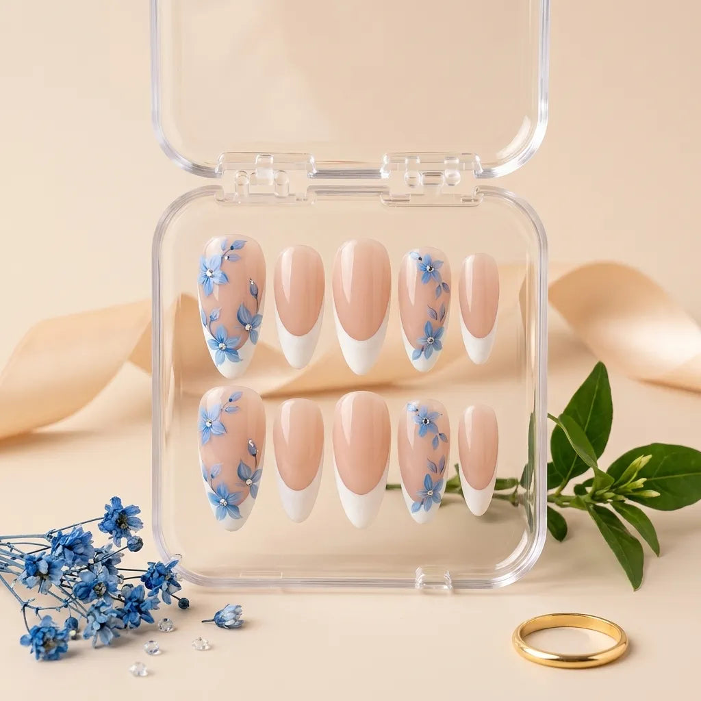 A set of false press-on nails displayed in a clear plastic case, featuring a nude pink and white French tip design with blue floral accents on two nails. The case is styled on a beige surface with decorative blue flowers, a green stem, and a gold ring.