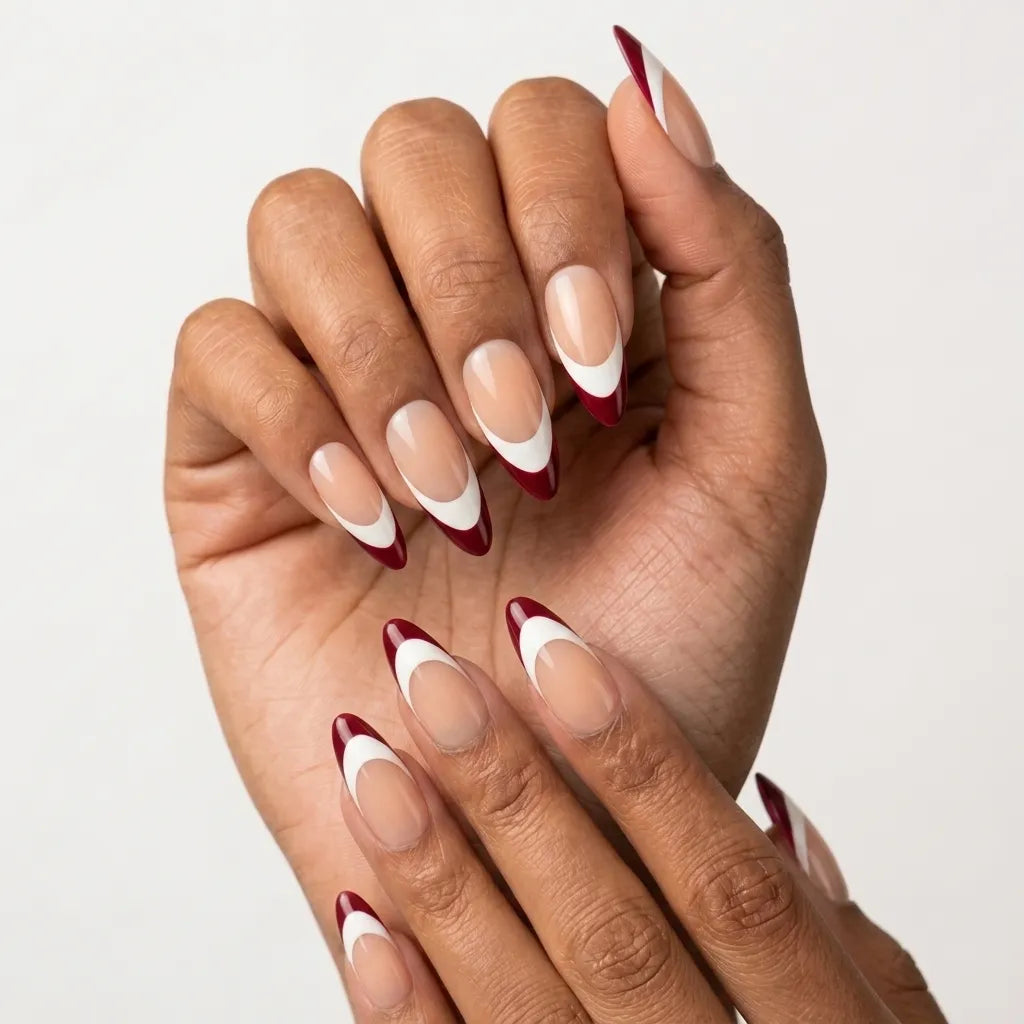 A set of eight false press-on nails in a clear plastic case, arranged on a beige background with dried rose petals, a dark leaf, and a white stone. The nails have a nude beige base with a white curved stripe and a deep red tip.