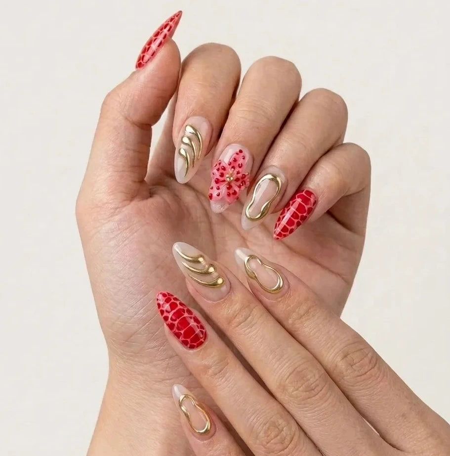 A close-up of hands showcasing a detailed almond-shaped nail art design. The manicure features a mix of solid red with a reptile pattern, red with a floral motif, and clear nails decorated with gold foil swirls and leaf designs, set against a plain light background.Handmade reusable press on nails