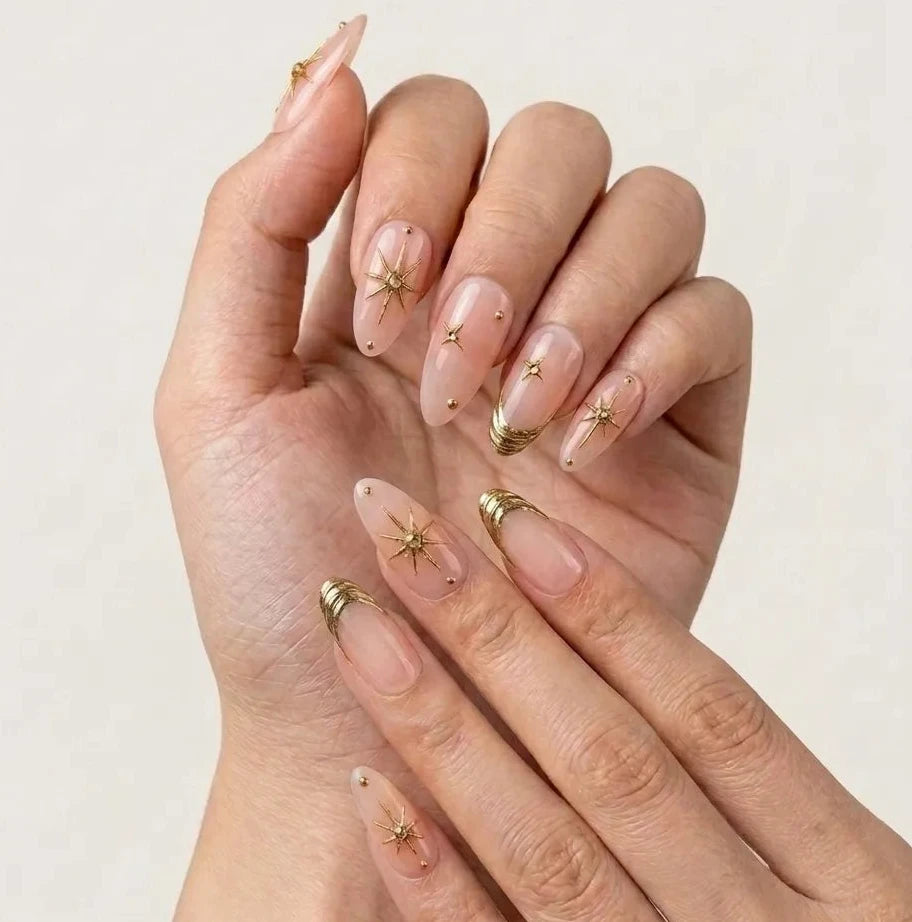 A close-up of hands showcasing a manicure with long, almond-shaped nude pink nails decorated with metallic gold starburst and crescent moon designs.Handmade reusable press on nails.Handmade reusable press on nails
