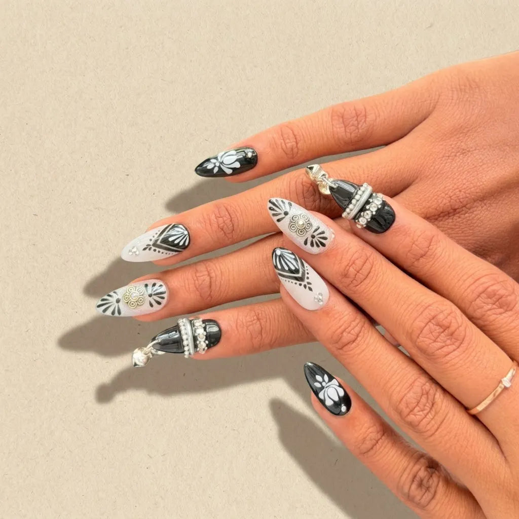 A close-up of a hand displaying a set of long, almond-shaped press-on nails with a black and white bohemian pattern, featuring floral motifs and silver rhinestone accents, against a neutral beige background.