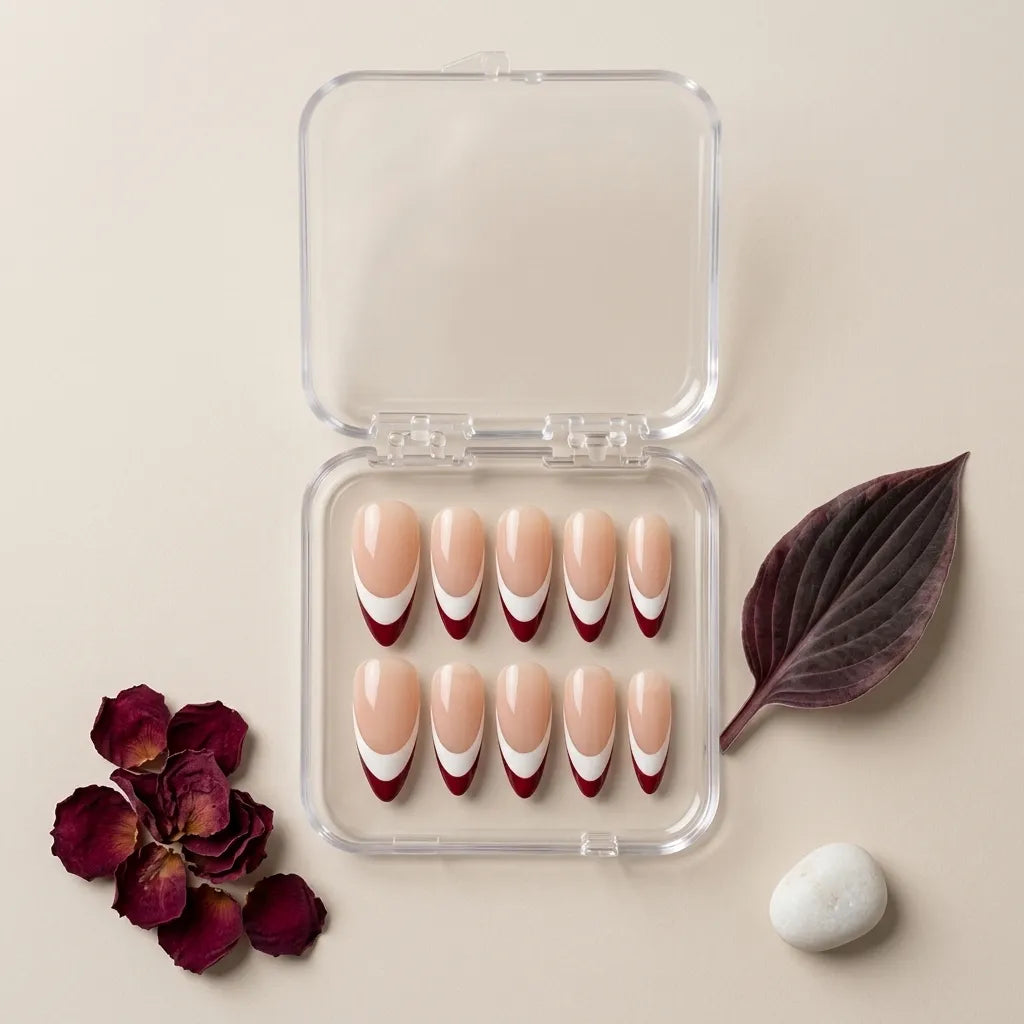 A set of eight false press-on nails in a clear plastic case, arranged on a beige background with dried rose petals, a dark leaf, and a white stone. The nails have a nude beige base with a white curved stripe and a deep red tip.