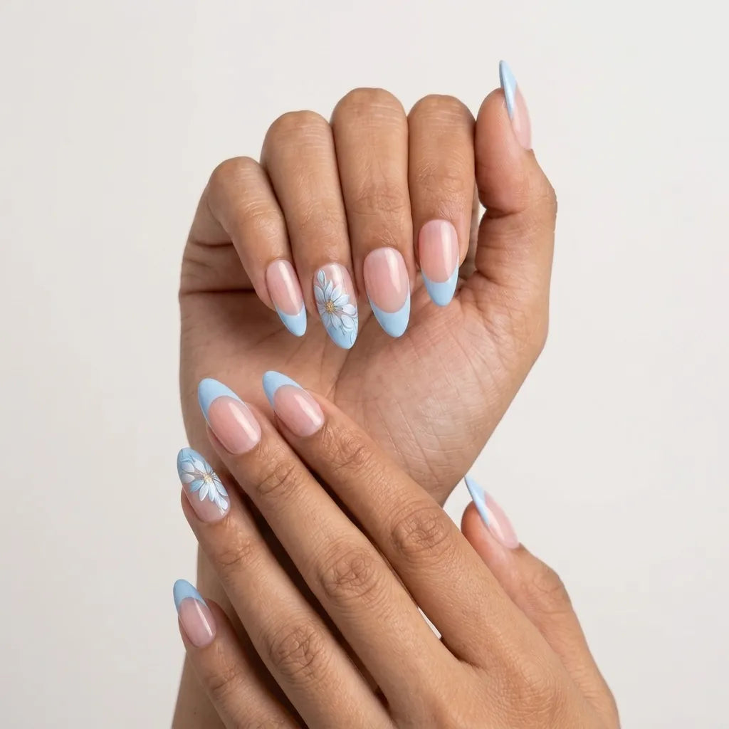 A set of ten false press-on nails in a clear plastic case, featuring a nude beige top half and a light blue bottom half, with one nail decorated with a small white flower. The case is styled on a green marble surface with decorative props.