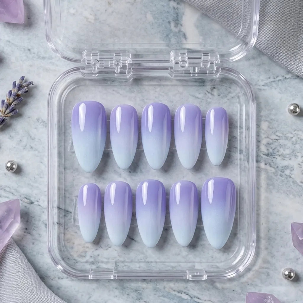 A set of ten false press-on nails with a purple-to-white ombre gradient, displayed in a clear plastic case on a light blue marble surface with lavender and crystal props.