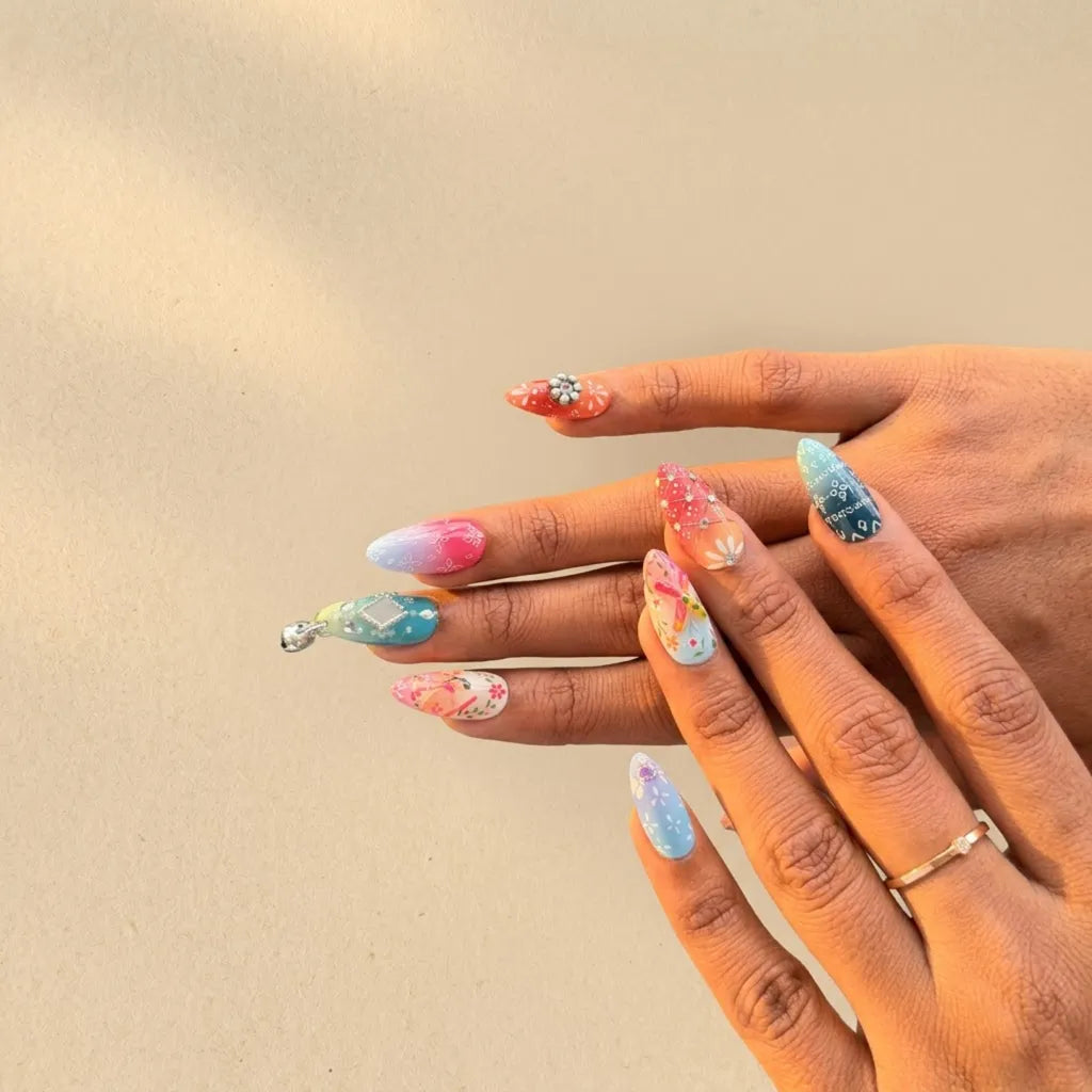 A close-up of a person's hands showcasing a variety of colorful, decorative press-on nails. The nails feature different designs including pastel watercolor gradients, glittery pink and blue patterns, floral prints, and one nail with a small silver rhinestone accent.