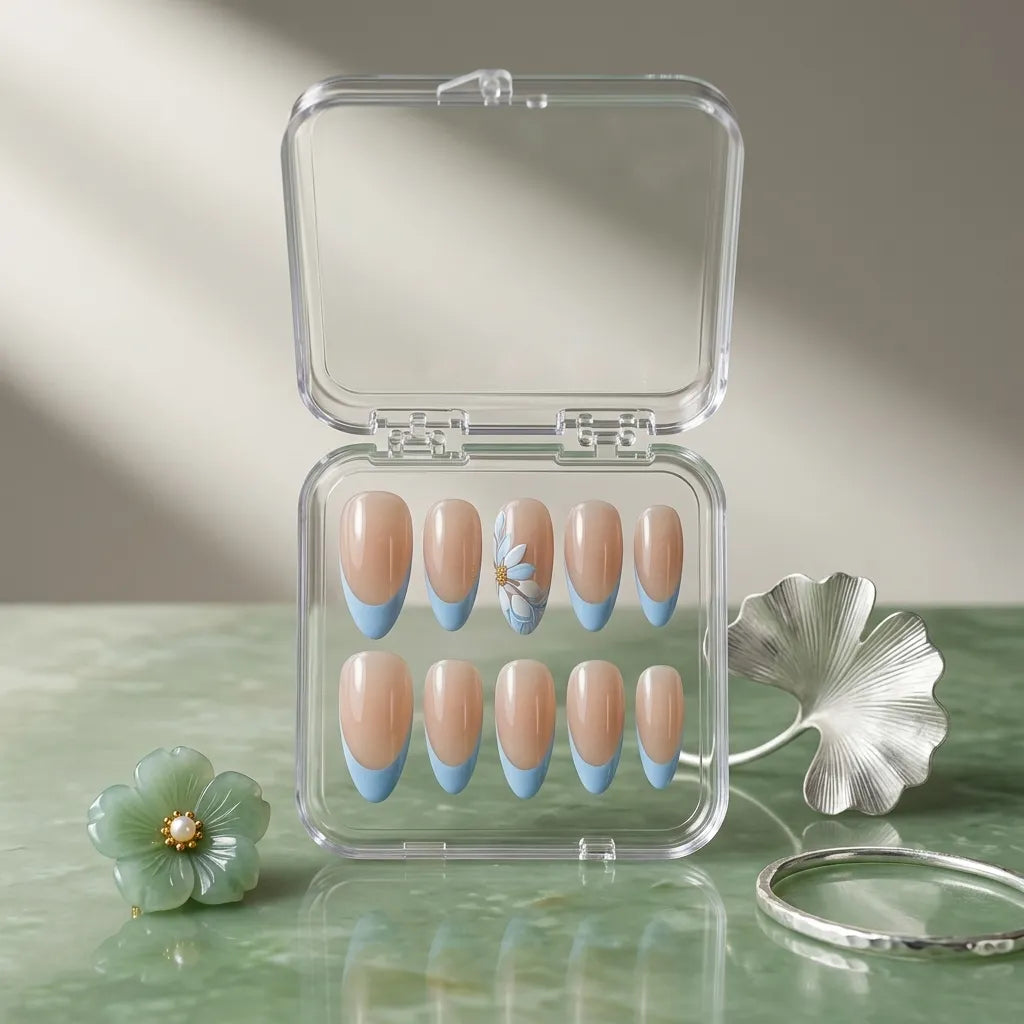 A set of ten false press-on nails in a clear plastic case, featuring a nude beige top half and a light blue bottom half, with one nail decorated with a small white flower. The case is styled on a green marble surface with decorative props.