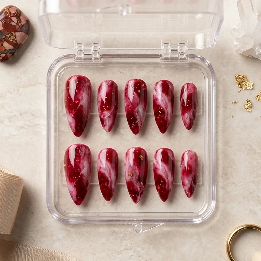 A set of ten false press-on nails with a deep red and white marble swirl pattern and tiny gold glitter flecks, displayed in a clear plastic case on a beige stone surface.