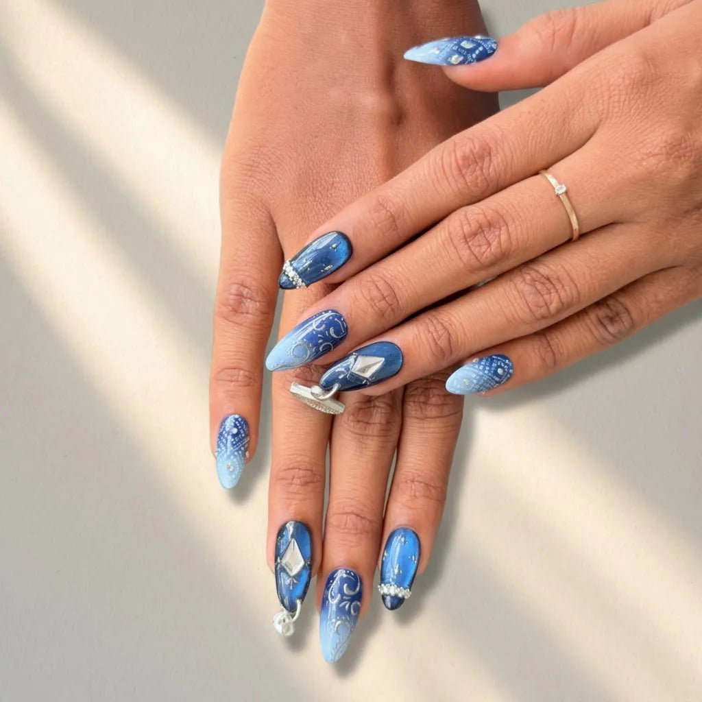 A close-up of hands showcasing a blue and white allover nail art design with intricate patterns and small rhinestone accents. The nails are shaped in a long almond style, and the hands are resting against a neutral light gray background with soft diagonal shadows.