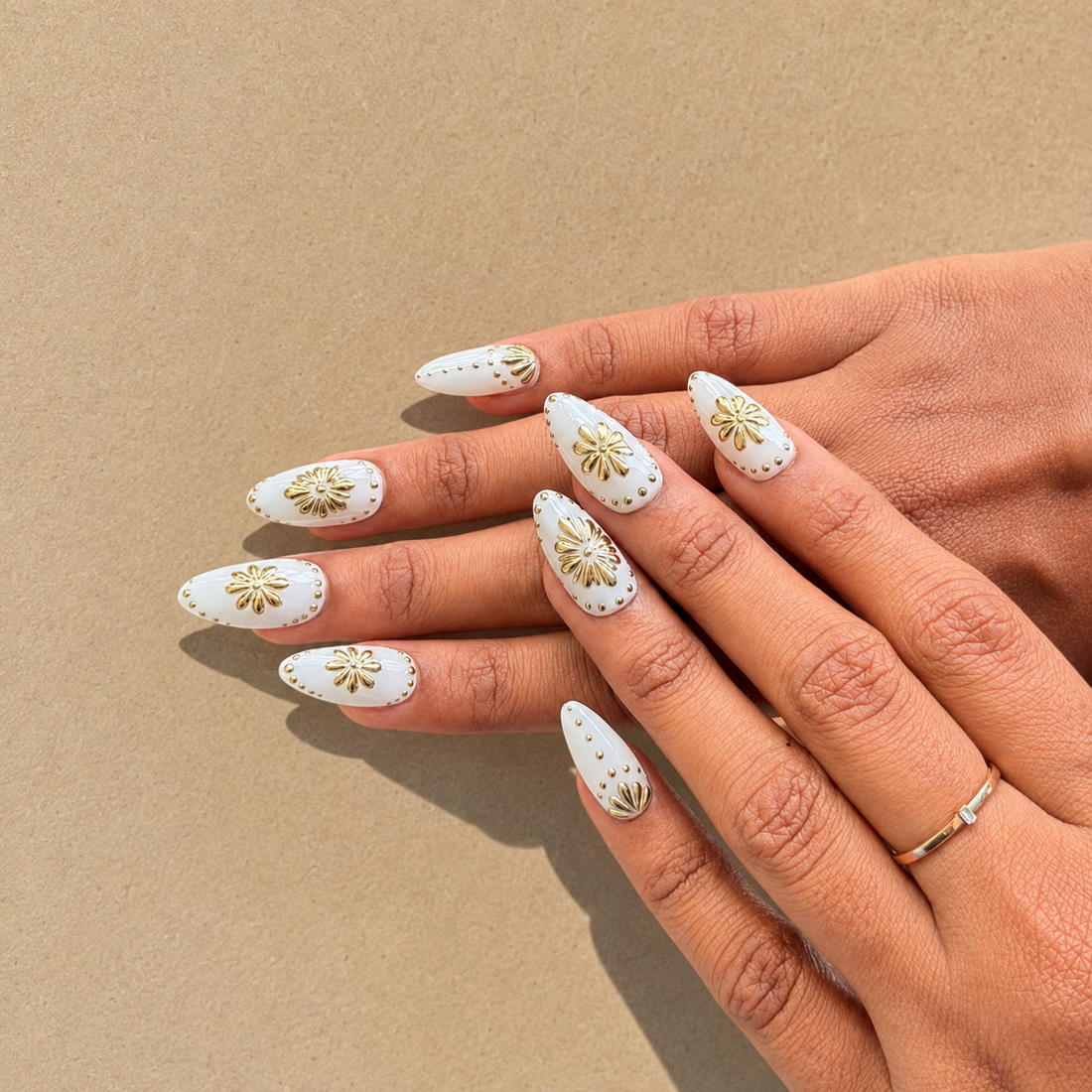 A close-up of a hand with long stiletto-shaped nails painted white and decorated with gold floral motifs and tiny gold dots, resting against a warm beige background.