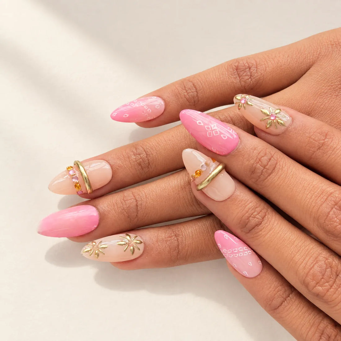 A close-up of a hand showcasing a set of long, almond-shaped press-on nails. The nails feature a mix of solid pastel pink, sheer nude pink, and clear bases decorated with delicate gold foil floral designs and small orange rhinestone accents.