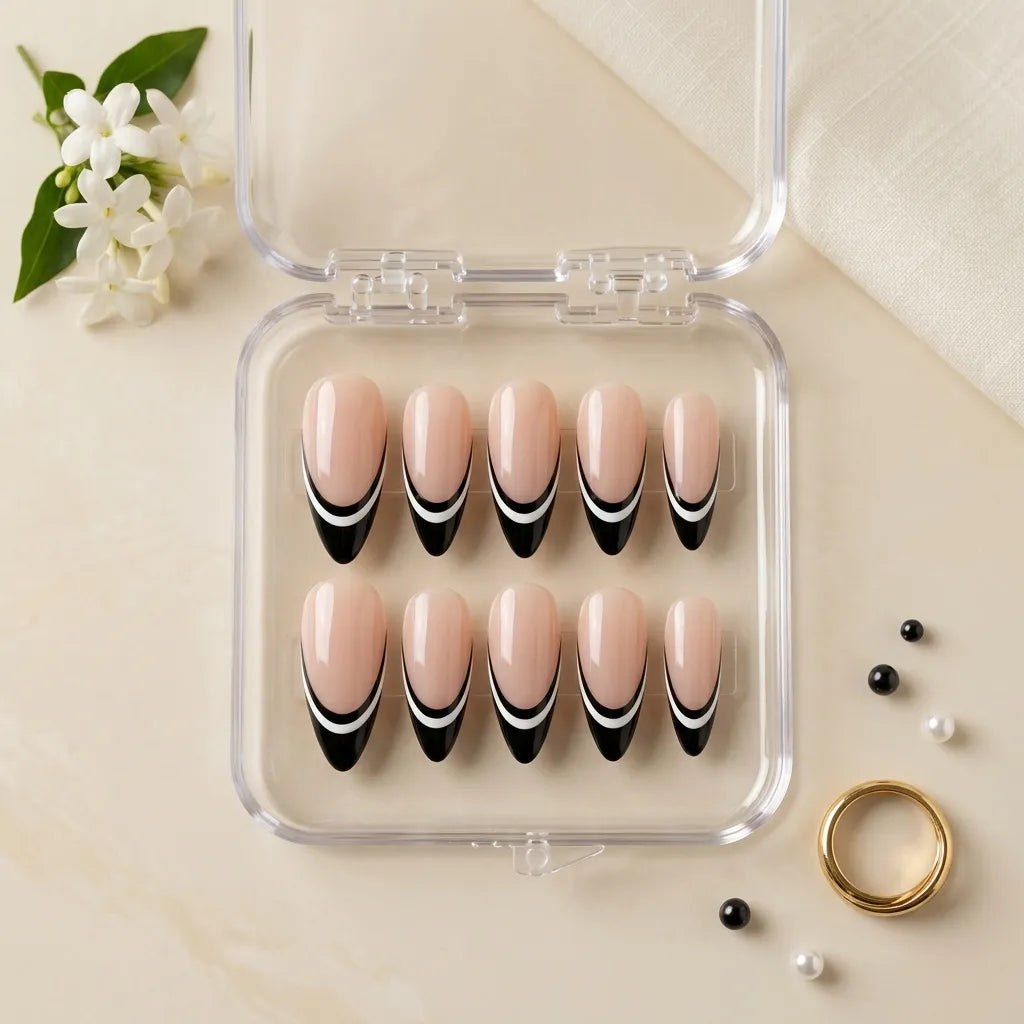A set of 10 false press-on nails in a clear plastic case, featuring a nude pink base with a black and white French tip design. The case is styled on a beige surface with small decorative props including white flowers, pearls, and a gold ring.
