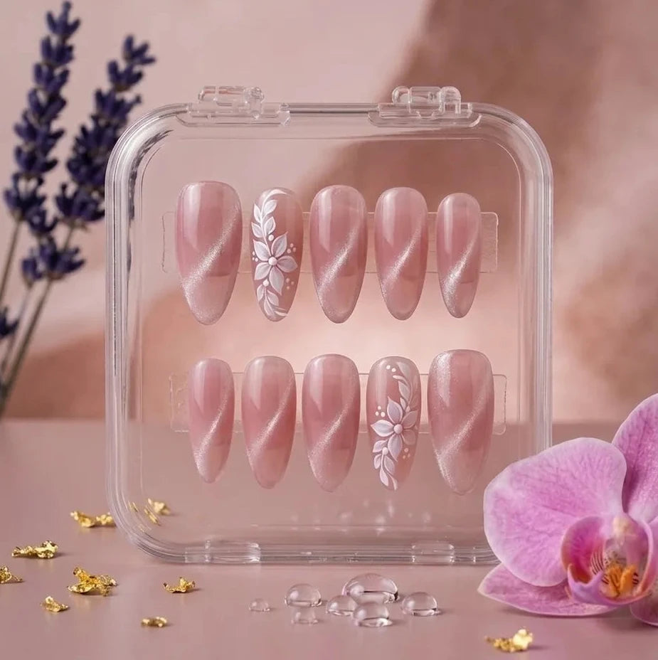 A set of 10 press-on nails in a clear plastic case, featuring a glossy nude-pink base with white floral designs on two nails, displayed on a pink surface with decorative lavender and an orchid flower.