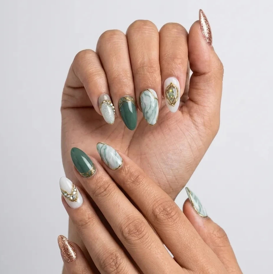 A close-up of two hands showcasing an elegant almond-shaped nail art design. The manicure features a mix of solid sage green, white with marble patterns, and white with gold crescent moon and gemstone designs, alongside metallic rose gold glitter tips.Handmade reusable press on nails