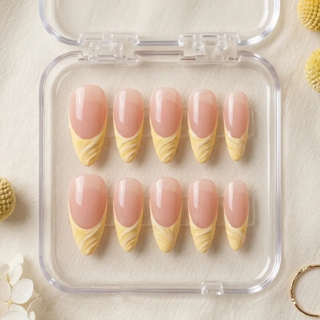 Ten false press-on nails arranged in a clear plastic case on a light beige fabric background, with decorative flowers and a gold ring around the edges. The nails have a glossy pale pink base with a yellow marbled pattern at the tips.