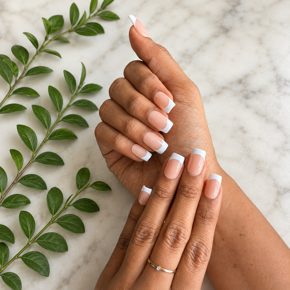 Ten press-on nails with a glossy nude pink and white French tip design, displayed inside a clear plastic case on a light beige textured background with subtle dried floral props.
