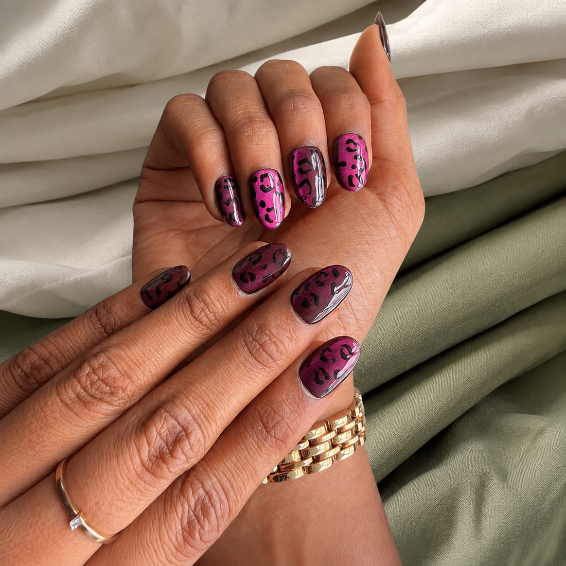 A set of ten false press-on nails with a dark burgundy base and black leopard spots, displayed inside a clear plastic case on a marble surface with decorative dried roses and gold flakes.