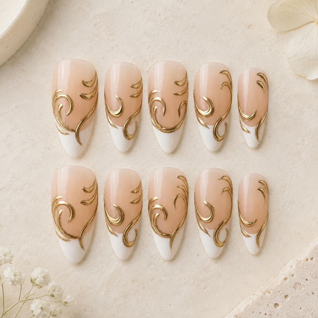 Ten almond-shaped press-on nails arranged in two rows on a light beige textured surface. The nails feature a glossy nude-peach upper half with a white lower half and elegant metallic gold scrollwork designs.