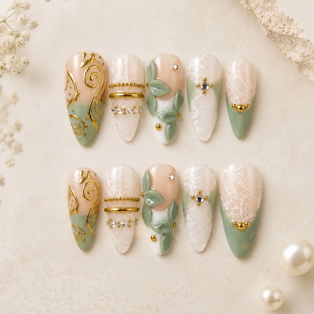 A set of 10 decorative press-on nails arranged in two rows on a light cream textured background with floral and lace props. The nails feature a mix of pale peach, mint green, white lace, gold foil swirls, pearl-like bead details, and small rhinestone accents.