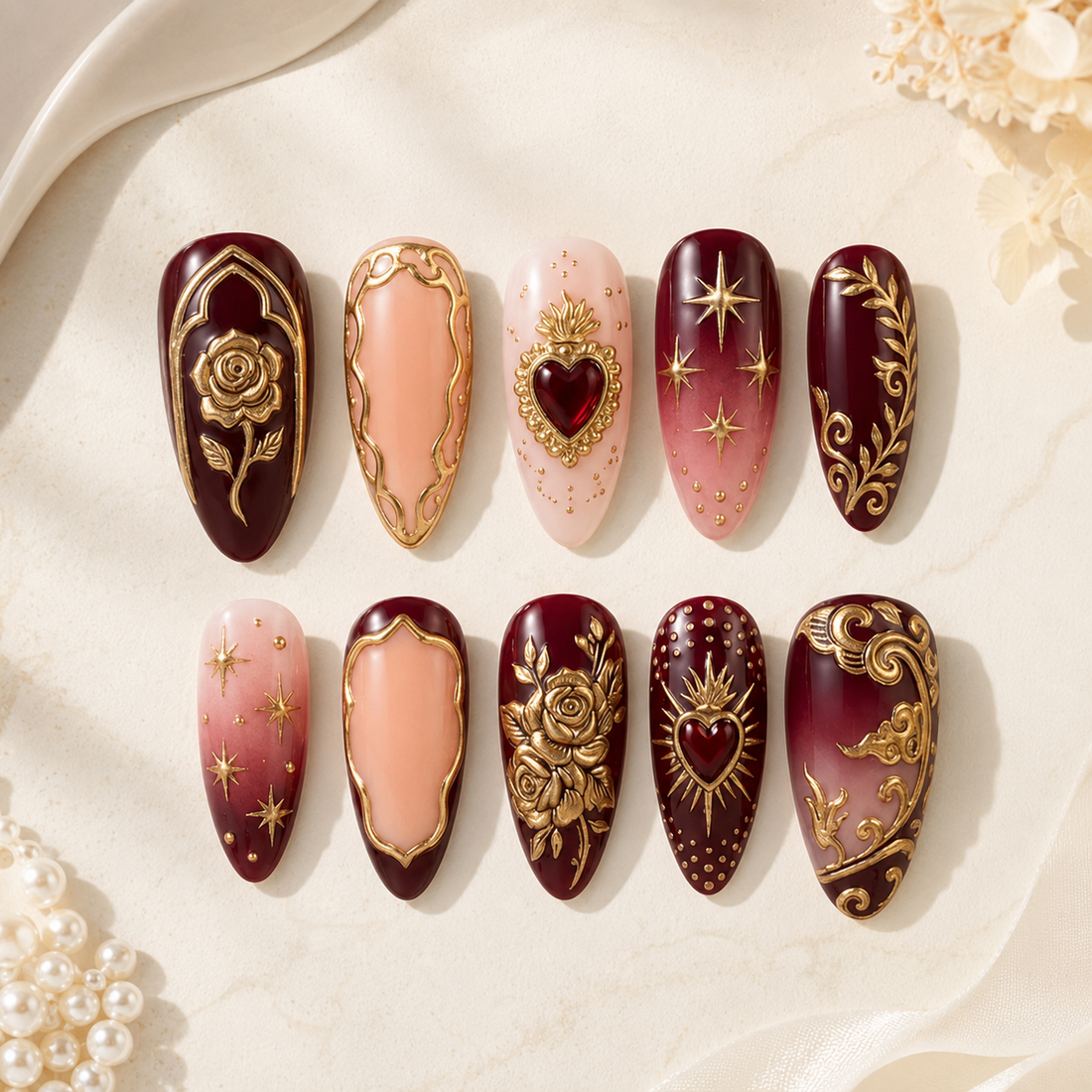A set of ten decorative press-on nails arranged in two rows on a light cream surface with soft fabric and pearl props. The nails feature deep burgundy, blush pink, and gold ornamental designs including roses, stars, hearts, and scrollwork.