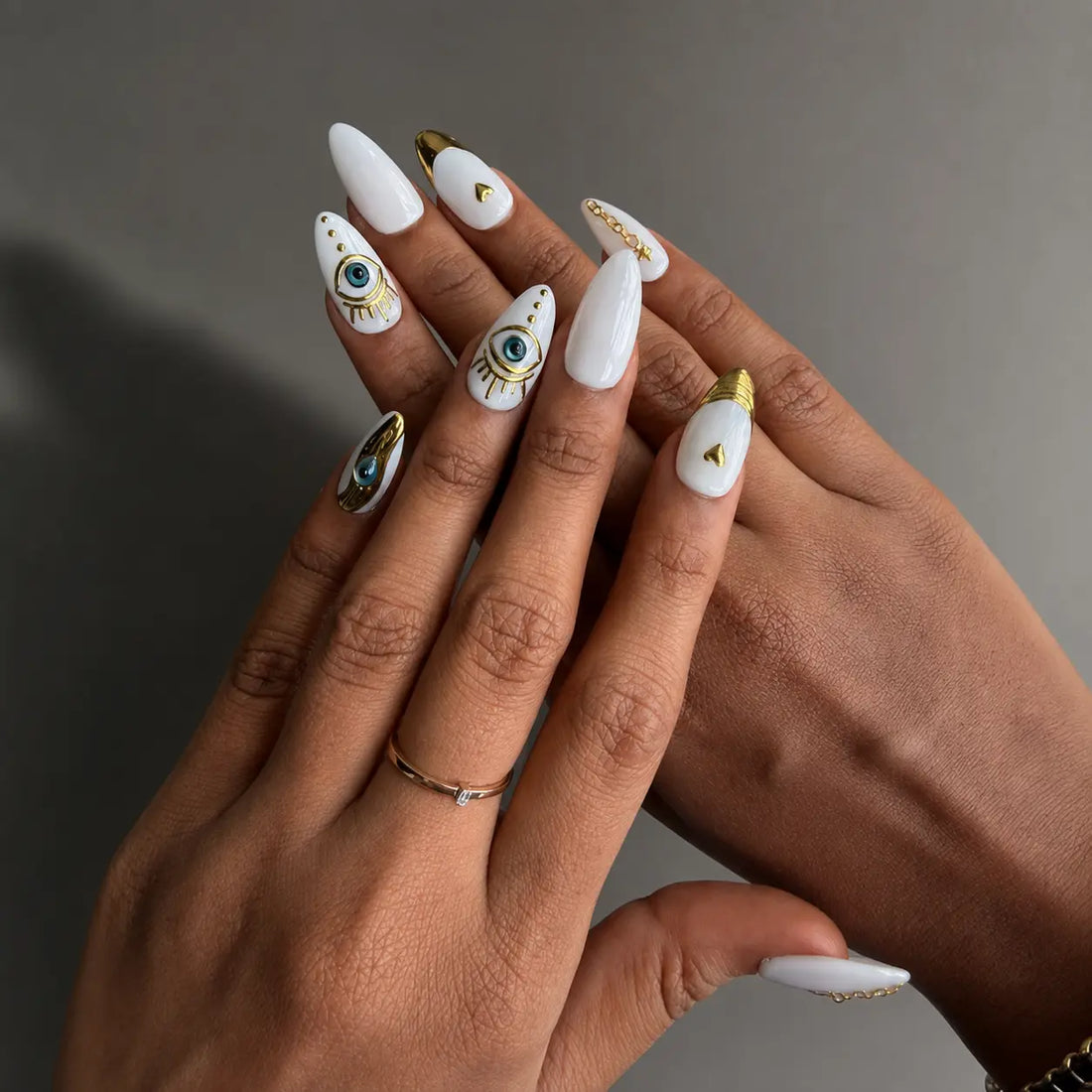 A set of decorative press-on nails displayed in a clear plastic case. The nails are primarily white and cream with gold metallic accents, including one blue eye design, one blue teardrop, and gold heart and star motifs.